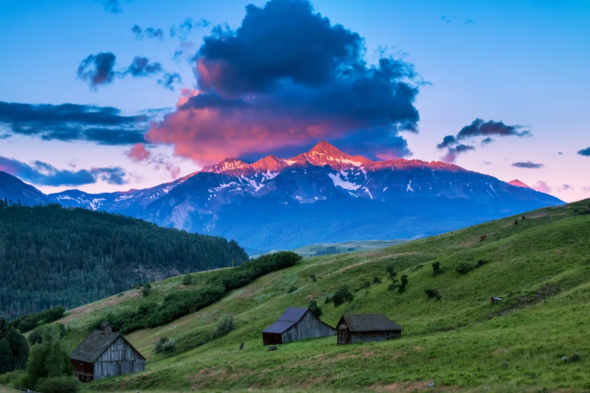 A sweeping view of the Rocky Mountains at sunset, with lush green hills in the foreground and rustic wooden cabins dotting the landscape. The sky glows with vibrant colors, while people can be seen hiking and relaxing, embracing the outdoors. The scene radiates tranquility and the awe of mountain adventure.