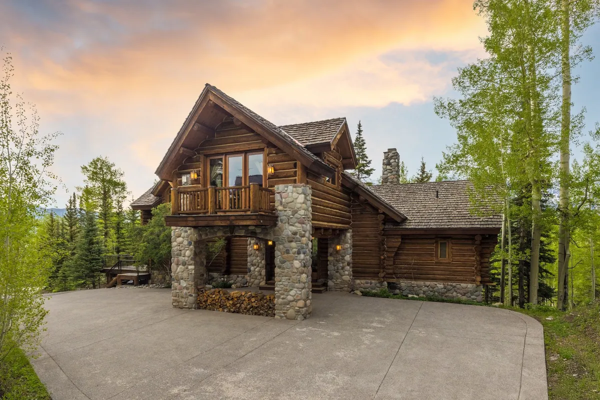 A rustic log cabin with a wooden balcony and stone pillars set against a sunset sky, surrounded by trees. The inviting outdoor space hints at family gatherings, outdoor relaxation, and enjoying the natural beauty year-round.