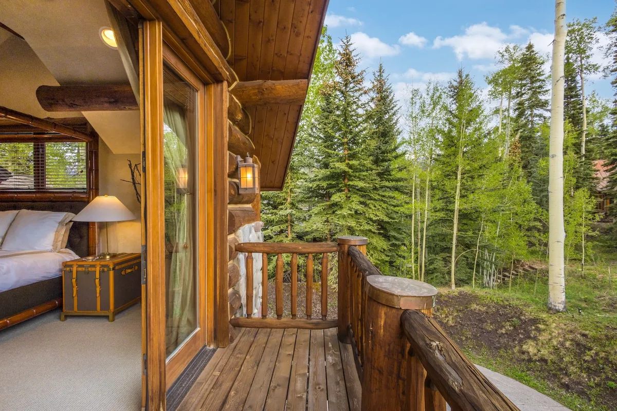 A rustic log cabin exterior with a wooden balcony overlooks lush green trees and natural surroundings. The scene is serene, with soft sunlight filtering through the foliage, and someone is relaxing on the balcony, enjoying the peacefulness and connection to nature.
