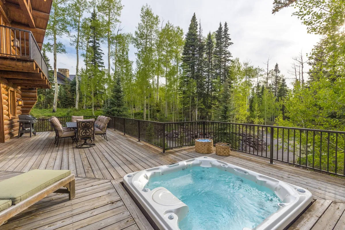 A serene wooden deck nestled among trees, featuring a large private hot tub with steam rising, comfortable outdoor seating, and a view of the natural surroundings. The space feels inviting and secluded, perfect for relaxation and social gatherings.