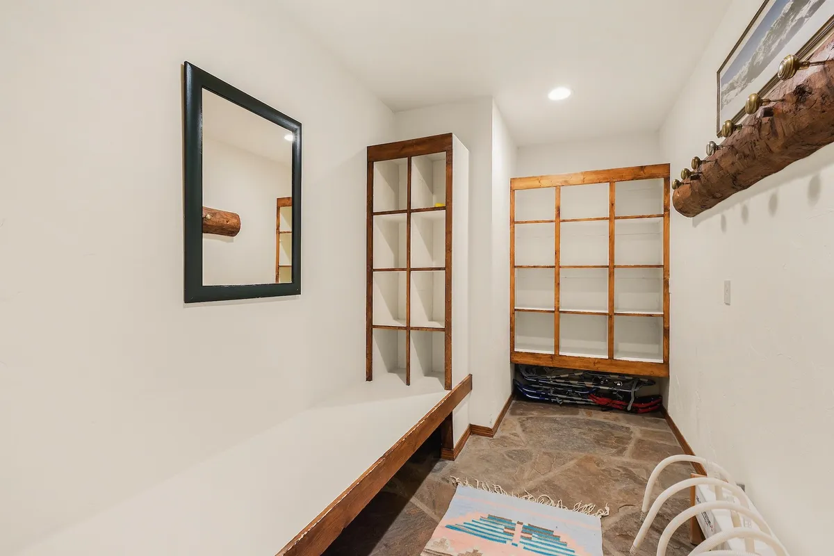 A welcoming entryway area featuring a sturdy wooden bench, a large mirror, stone flooring, and a wooden shelving unit with ample space for storing outdoor gear. The decorative rug and casual, functional layout suggest it's tailor-made for guests transitioning from the outdoors, organizing boots, and storing winter or hiking gear.