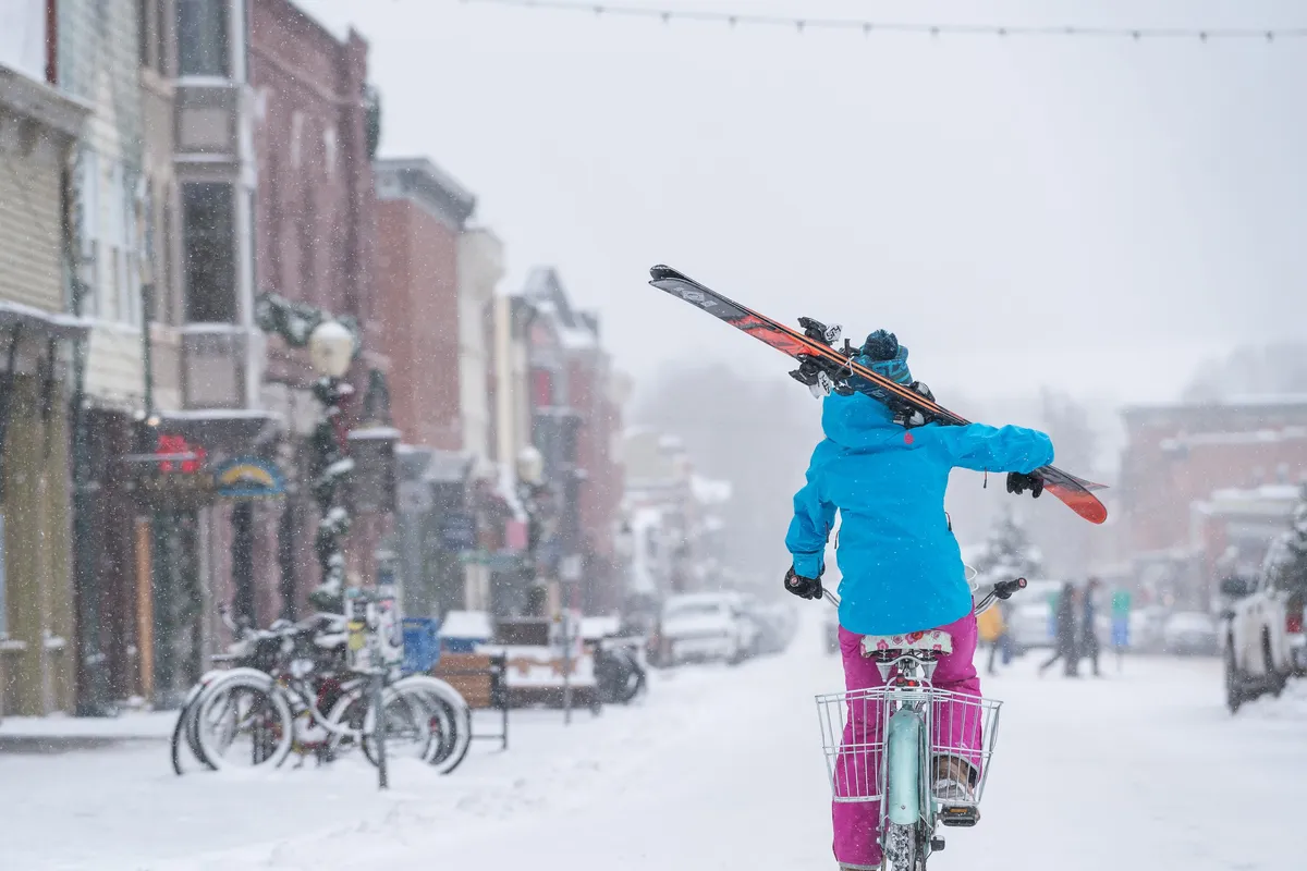 A person carrying skis walks along a snow-covered street in a lively village area, with bicycles, quaint buildings, and the vibrant atmosphere of a mountain town. The scene hints at the convenience of lifts, local shops, and shuttle access.