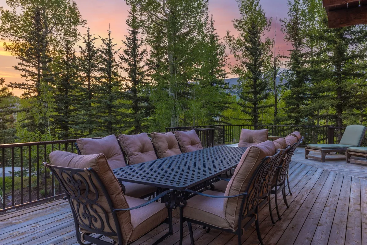 A spacious outdoor dining area features a large wooden table surrounded by comfortable seating, set amidst lush trees with a glowing sunset sky in the background. The welcoming arrangement invites a group to share a meal, converse, or celebrate together outdoors.