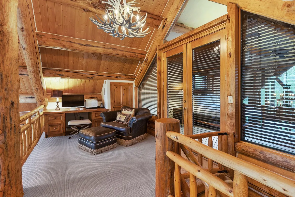 A warm, rustic office nook features a wooden desk, leather armchair, antler chandelier, and large windows with blinds. The space is ideal for working, reading, or quiet reflection, with a cozy and inviting mood.