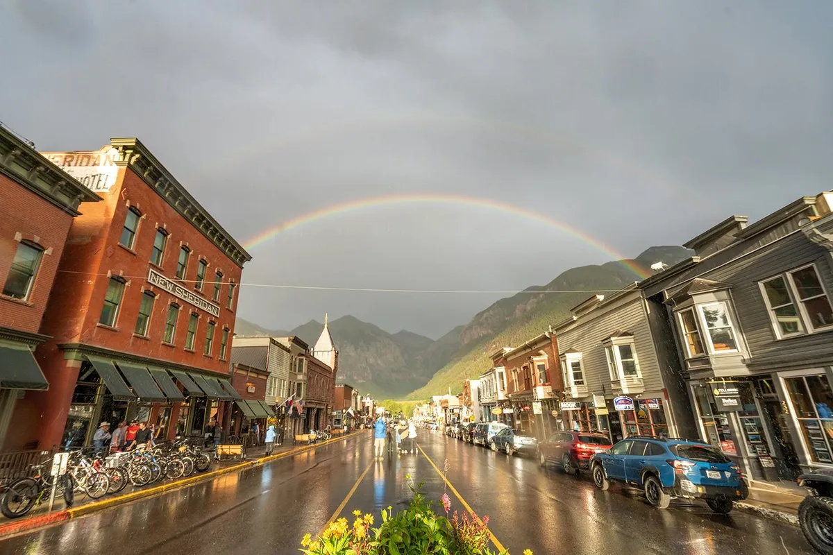 A charming street scene featuring historic buildings, a vivid rainbow stretching across the sky, and reflections in the wet pavement. Bicycles are parked along the sidewalk, and the area feels lively yet upscale, hinting at the variety of dining and shopping options nearby Mountain Village.