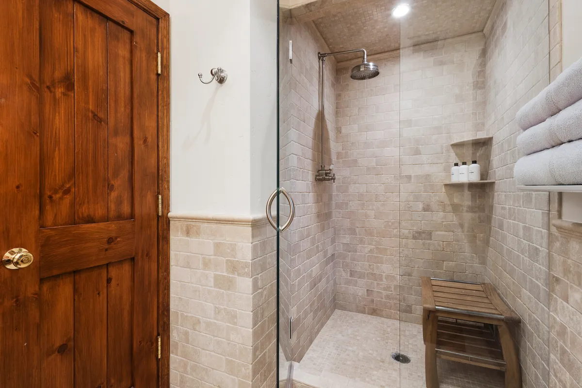 A tranquil bathroom with a glass-enclosed, stone-tiled shower, wooden bench, and neatly stacked towels. The inviting ambiance and practical design evoke a luxurious post-adventure refresh, ideal for unwinding after a day on the slopes or trails.