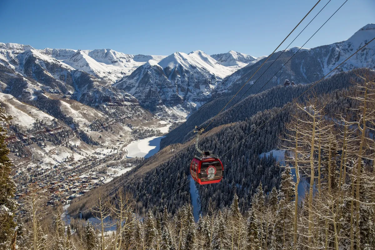 A sweeping view from the gondola looking out over snow-covered mountains, a picturesque valley, and the town below. The gondola cabins are visible suspended above the landscape, symbolizing easy access to the Mountain Village and its scenic surroundings.