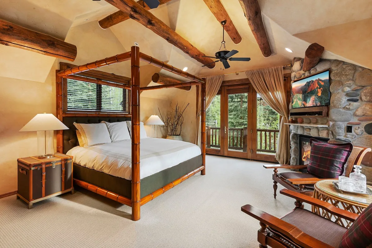This interior image showcases a four-poster king bed in a spacious bedroom, with a stone fireplace, large windows offering views of nature, and a comfortable seating area. The ambiance is tranquil and luxurious, emphasizing privacy and relaxation.