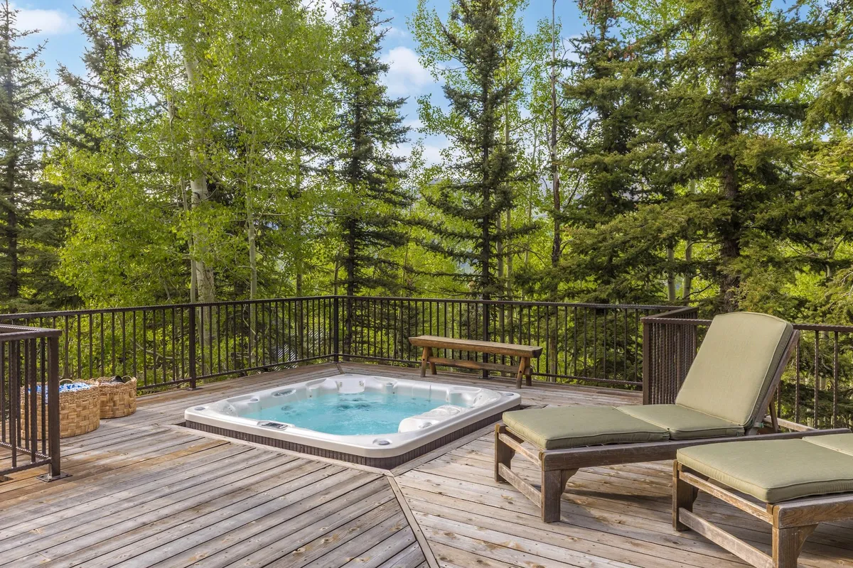 A stylish wooden deck furnished with lounge chairs and a bubbling hot tub, surrounded by leafy trees. The scene captures the warmth of sunset, inviting guests to unwind and soak in the views.