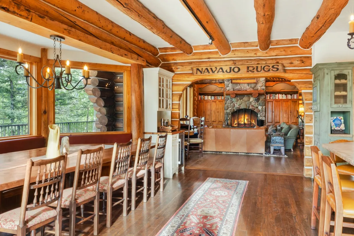 This dining area features a long wooden dining table with custom bench seating next to a cozy fireplace, under rustic wooden beams. The space is designed for group feasts, games, and memorable storytelling, capturing the welcoming and communal spirit of the cabin.