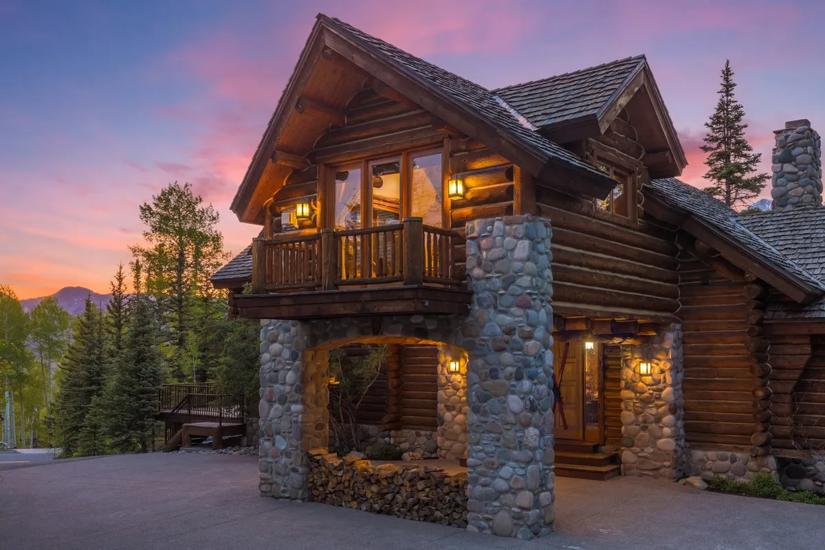 The image showcases a stunning modern log cabin with robust stone pillars, expansive windows, and a spacious balcony framed by a rustic railing. The home is set against a peaceful mountain backdrop, inviting gathering spaces for families or friends to enjoy sunsets or fireside chats outdoors.