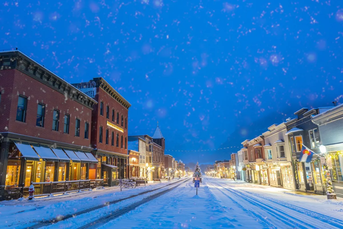 A charming snow-covered village street lined with decorated shops, glowing street lights, and a festive Christmas tree. The ambiance is warm and inviting, with hints of holiday activities and a lively yet cozy atmosphere.