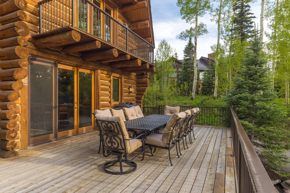 The log cabin’s spacious outdoor dining area is set for a meal, surrounded by tall green trees. The deck invites gatherings for grill-outs or evening stargazing, with warm lighting hinting at coziness and relaxation in nature.