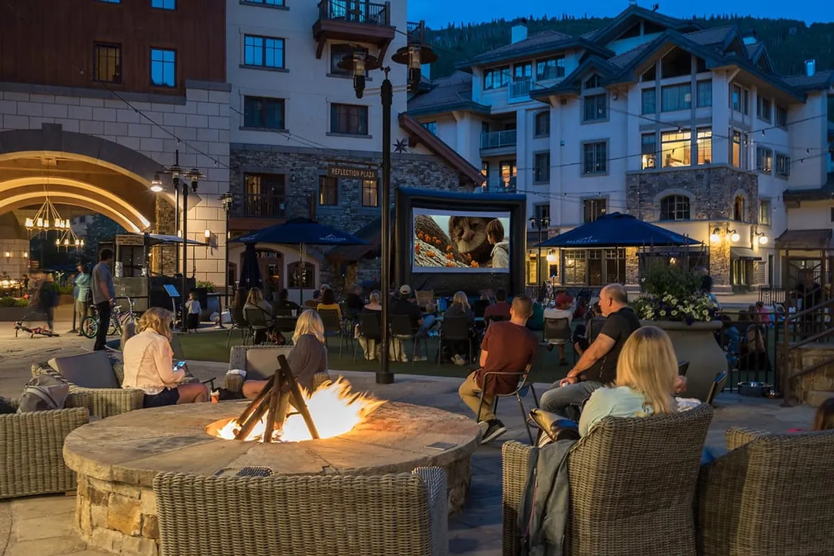 A cozy exterior setup with a fire pit, tiered seating, and an outdoor movie screen, surrounded by architectural details of the cabin. The scene is lively yet intimate, conveying a sense of community and relaxed enjoyment during an evening movie or fireside gathering.