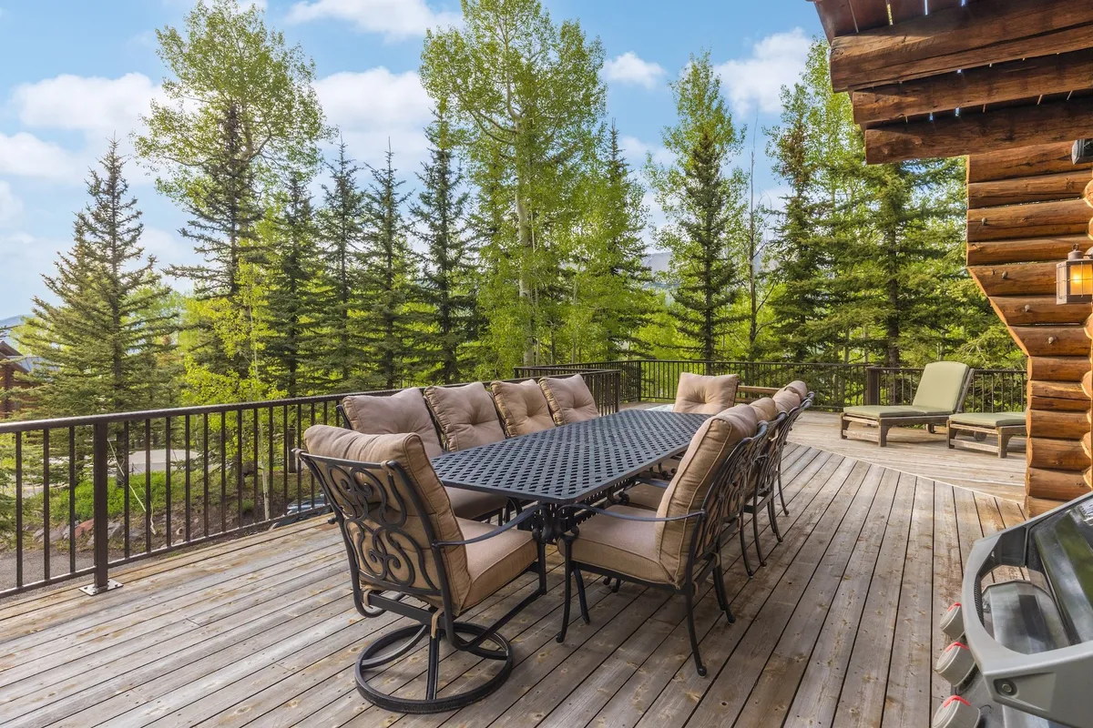 An expansive outdoor deck with a large dining table set for al fresco meals, surrounded by trees and nature. Lounge chairs offer a place to unwind, and the deck railing frames the scenic outdoor setting.