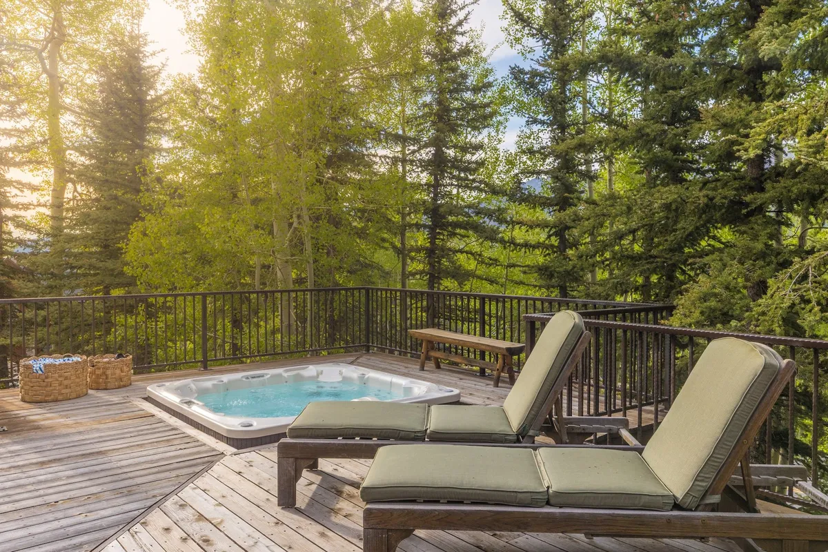 A steaming hot tub sits on a deck nestled among towering trees, with comfortable lounge chairs nearby. The tranquil setting suggests relaxation and après-ski enjoyment, surrounded by the beauty and peace of nature.