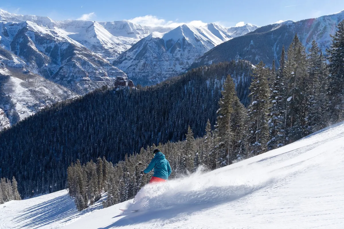 A skier in motion glides down a snowy slope surrounded by snow-covered mountains and evergreen trees. The action shot captures the excitement and beauty of the Telluride ski experience, with crisp mountain air and pristine powder on full display.