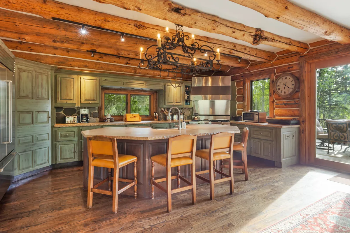 The kitchen is showcased here with a large central island perfect for casual gatherings, an ornate chandelier above, green-finished custom cabinets, and high-end appliances. Large windows ensure the space is filled with natural light, and the layout encourages family interaction.