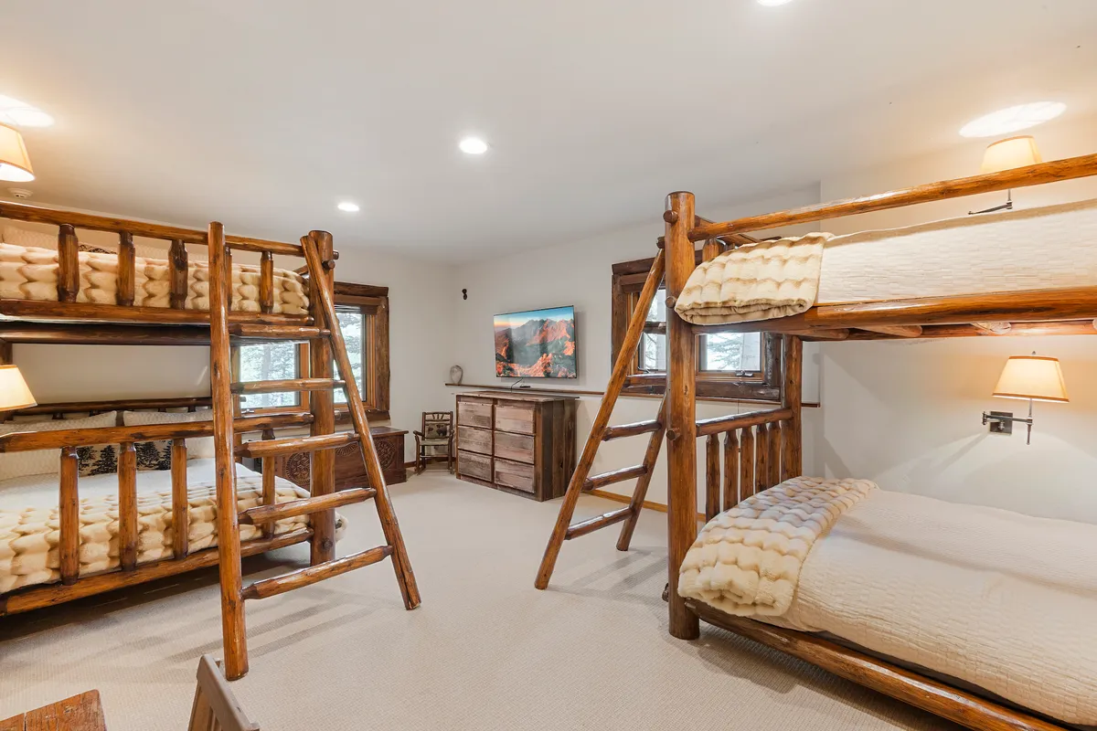 A playful and inviting bunk room with multiple bunk beds, wooden furniture, a television, and windows letting in natural light. The space feels warm and welcoming, perfect for groups or families.